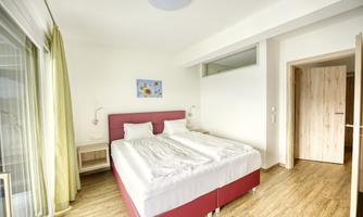 A modern bedroom with a large double bed and wooden floor. The walls are bright, and there is a window with curtains.