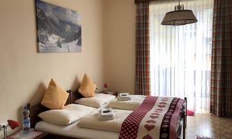 Cozy bedroom with two beds and decorative pillows. The room has a window with curtains and a picture of a mountain landscape on the wall.