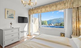 A bright bedroom with a window overlooking a picturesque mountain landscape. There is a bed, a television, and an elegant ceiling lamp.