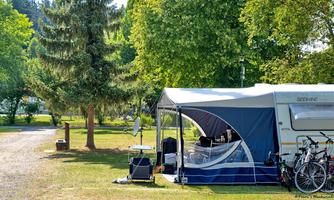 A caravan with an awning is located in a green camping area. Next to the caravan, there are bicycles and trees surround the site.