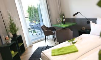 A modern room with a bed, a seating area, and a small table. Large windows provide plenty of light and lead to a balcony with a view of the greenery.