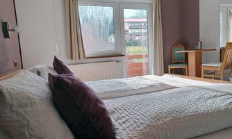 A cozy room with a double bed and large windows. The view looks out onto a quiet outdoor area with chairs and a table.