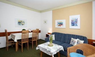 A bright and welcoming living room with a blue sofa and a dining table. The walls are decorated with colorful pictures, and the furniture is simple and cozy.