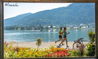 Two joggers by the shore of a picturesque lake, surrounded by flowers and green mountains. In the background, a charming cityscape is visible.