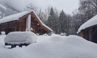 A snowy landscape with wooden chalets and snow-covered trees. The sky is cloudy, and the surroundings feel calm and wintry.