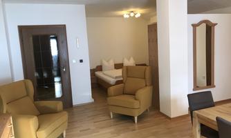 A cozy apartment with two comfortable armchairs, a dining table, and a double bed in the background. The room has a bright wooden floor and a friendly atmosphere.