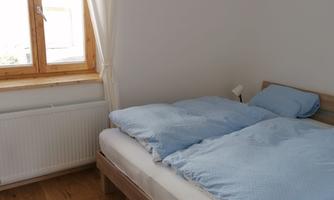 A cozy bedroom with a double bed and blue bedspreads. The room has a large window that lets in plenty of daylight.