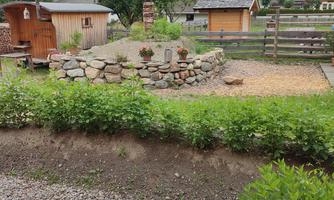 A picturesque garden with stones, surrounded by green plants and a view of the mountains. Cozy huts and a fence can be seen in the background.