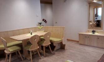A cozy dining area with a wooden table and four chairs in a bright room. The walls are plain, and there is a large mirror dresser.