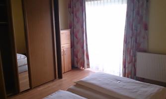 A bright bedroom with two beds and a large window. The room is decorated with a wardrobe and curtains.