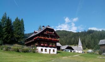 A beautiful old house surrounded by green meadows and trees. In the background, mountains and a clear blue sky can be seen.