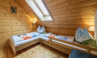 A cozy attic room with wooden paneling and two beds. Large windows provide plenty of daylight.