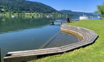 A calm lake with green shores and a wooden pier. In the background, gentle hills and boats can be seen.