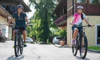 Two people are riding their bicycles on a picturesque street. Surrounded by beautiful buildings and lush nature, they are enjoying a sunny day.