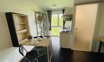 A modern kitchen corner with a dining table and two chairs. The room has large windows with a view of greenery.