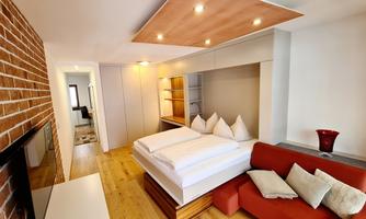 A modern bedroom with a cozy bed, red sofa, and wooden floor. The walls are bright and designed with a touch of brick.