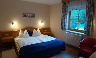 A cozy bedroom with a double bed and colorful pillows. The room has a window with curtains and a red armchair.