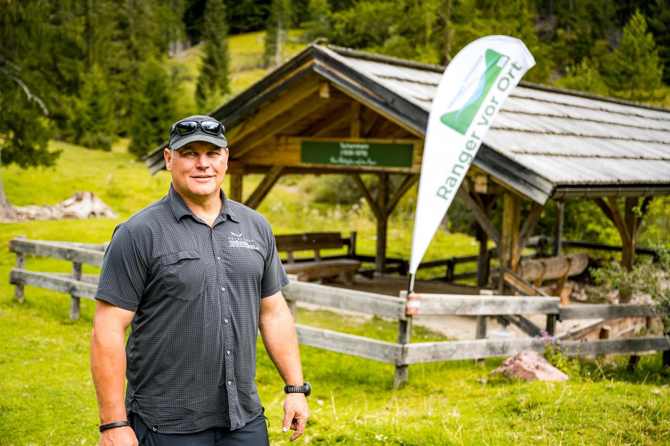 Ranger vor Ort: Mann mit Kappe und Sonnenbrille vor Holzunterstand in Bergregion
