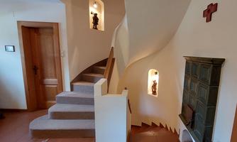 A bright staircase with a curved railing leads upwards. There are alcoves with small figures on the walls and a fireplace in the corner.