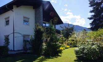 A beautiful house with a large garden and abundant green plants. In the background, the mountains and a clear blue sky are visible.