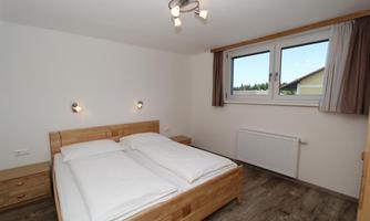 A simple bedroom with a double bed and wooden furniture. The walls are light, and there is a large window that lets in plenty of daylight.
