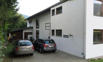 A modern residential house with large windows and a low-maintenance exterior. Two cars are parked in front of the entrance on a paved path.
