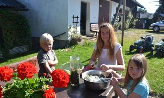 A woman and two children are sitting outside at a table preparing something. In the background, red geraniums are blooming and some gardening tools are visible.