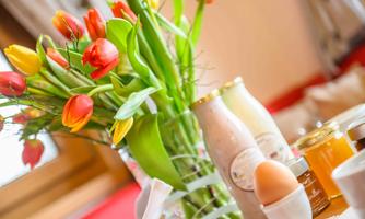 A colorful bouquet of tulips is standing in a vase on the table. Next to the flowers are various foods like jam, an egg, and bottles.