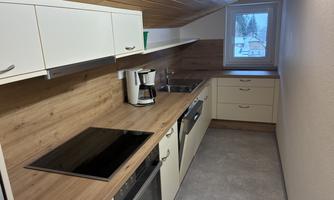 A modern kitchen with bright furniture and a wooden countertop. It features cooking fields, a sink, and a window with a view outside.