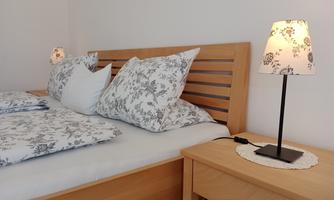 A cozy bedroom with a wooden bed, on which white bedding with a floral pattern lies. Next to the bed, bedside lamps are placed on a wooden nightstand.