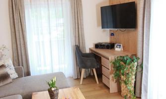 A cozy living room with a sofa, a wooden table, and a desk. The room is bright and friendly with a television and a small plant.
