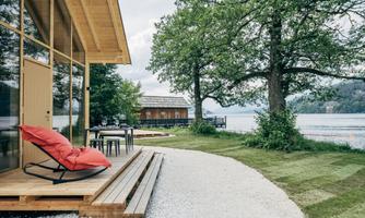 A modern wooden house by the lake with a terrace and a red lounge chair. Surrounded by green trees and a grassy meadow.