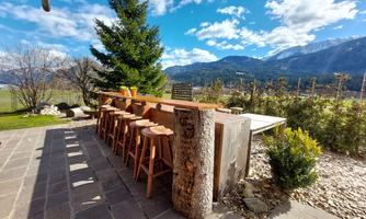 A cozy terrace with a wooden bar and stools. In the background, mountains and a blue sky can be seen.