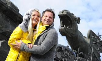 A happy couple stands in front of an impressive statue. They are wearing bright, warm clothes and smiling at the camera.
