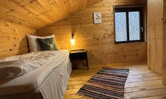 A cozy bedroom with wood paneling and a single bed. Soft light falls through the window and the floor is made of wood with a colorful rug.