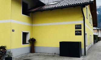 A yellow house with a pitched roof and a simple facade. The entrance is surrounded by a small plant, and the street runs straight past.