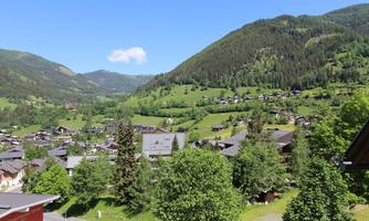 A picturesque landscape with green hills and a clear blue sky. In the foreground, houses and trees are visible.