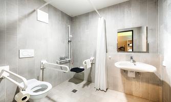 A modern, barrier-free bathroom with gray tiles. It includes a toilet with grab bars, a sink, and a shower area with a curtain.