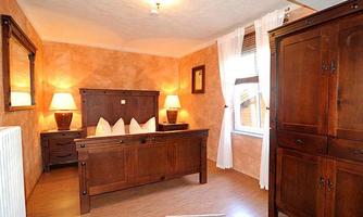 A cozy bedroom with wooden furniture and warm, earthy colors. There is a large bed, two nightstands, and windows with white curtains.