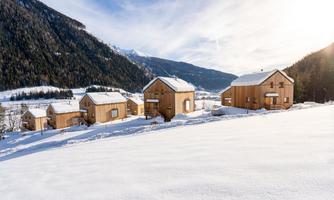 A picturesque winter landscape with modern wooden houses. The surroundings are covered in snow and surrounded by mountains.