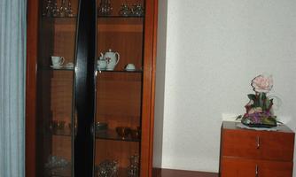 A wooden cabinet with glass doors that displays various decorative items. Next to it stands a small chest of drawers with a flower on top.