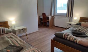 A bright room with two beds, nightstands, and a desk area. The windows let in plenty of daylight.
