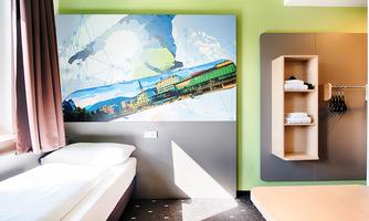 A modern hotel room with a bright mural and a simple sleeping area. The walls are designed in vibrant colors, and there is a shelf with towels.