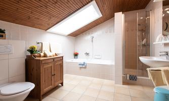 A modern bathroom with wooden flooring and a sloped ceiling. It contains a bathtub, a shower, and a sink, as well as stylish decorations.
