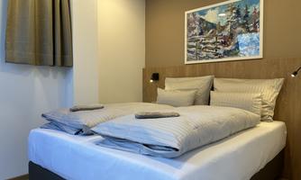 A modern bedroom with a large double bed and elegant bedding. A decorative picture hangs on the wall, and the windows are covered with curtains.