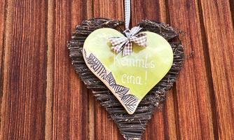 A heart-shaped wooden sign hangs on a wall. It has green writing that says: "Are you coming!" and it has a bow made of checkered fabric.