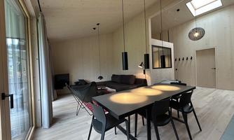 A modern living room with light wood interior. The room is furnished with a dining table, chairs, and a cozy seating area.