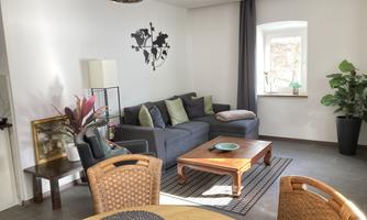 A cozy living room with a gray couch and a wooden table. The room is brightly lit and has plants as well as decorative elements.