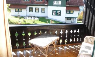 A balcony with a small table and chairs that offers a beautiful view of a two-story, traditional German house. In the background, well-kept meadows and decorative elements can be seen.