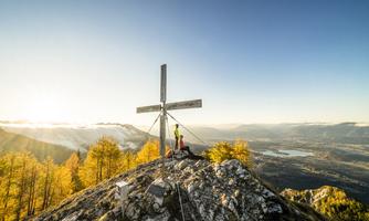 A mountain peak with a cross and a beautiful view of the valley. The sky is clear and the landscape features autumn-colored trees.